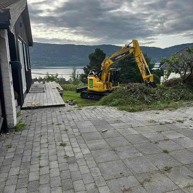 Gul gravemaskin rydder ved hus med utsikt over vann og fjell.